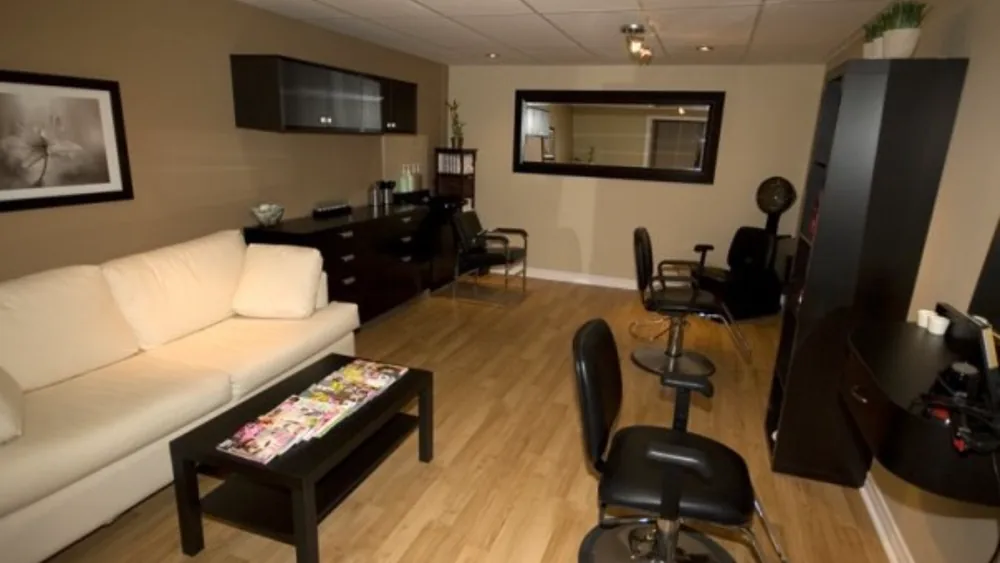 The salon room: a cream sofa and coffee table, two black styling chairs and a large mirror on a taupe wall.